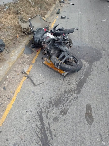 Una motocicleta negra destruida yace volcada en el borde de una vía pavimentada, rodeada de piezas rotas y manchas sobre el asfalto