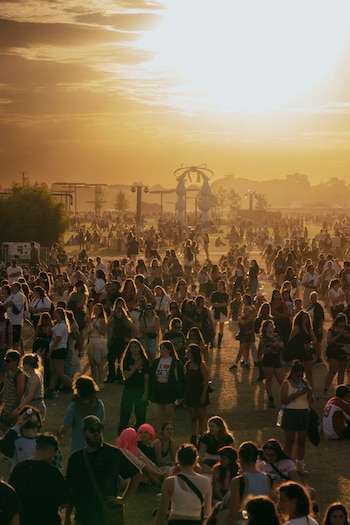 Vista aérea de una gran multitud de personas en un festival al aire libre al atardecer, con el sol bajo y un cielo teñido de tonos dorados