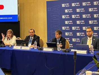 Cuatro personas sentadas ante una mesa con mantel azul, micrófonos, computadoras y un panel de fondo con logotipos institucionales