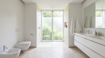 Un baño moderno y luminoso con ducha abierta sin mampara, inodoro, bidé, lavabo doble con espejo y dos toallas blancas. Una ventana grande ilumina la estancia.