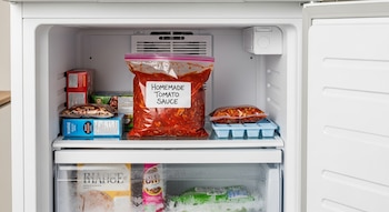 Congelador doméstico blanco abierto con una bolsa de salsa de tomate casera etiquetada, envases de comida congelada y una cubetera con porciones de salsa.