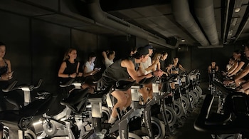 Vista lateral de un grupo de personas, hombres y mujeres, pedaleando intensamente en bicicletas estáticas en una sala de spinning con poca luz.