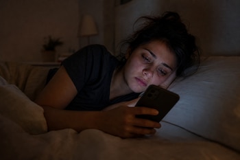 Una mujer recostada en una cama sostiene y mira la pantalla encendida de un teléfono celular en un entorno oscuro.