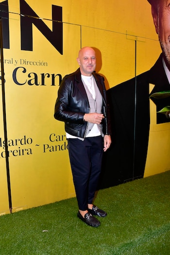 Hombre calvo con barba corta, viste chaqueta de cuero negra y pantalón oscuro, posa frente a un fondo amarillo con texto y un suelo de césped artificial