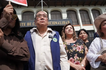 Primer plano de un hombre asiático mayor con gafas y chaleco azul, junto a varias personas, incluyendo una mujer con blusa floral, en un evento al aire libre