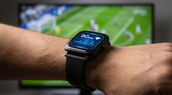 Primer plano de un brazo con un reloj inteligente negro mostrando 90 ppm y un gráfico de frecuencia cardíaca en una pantalla azul, con un partido de fútbol borroso de fondo.