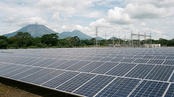 Campo de paneles solares en El Salvador bajo un cielo nublado, con grandes volcanes y vegetación tropical al fondo, junto a una subestación eléctrica.