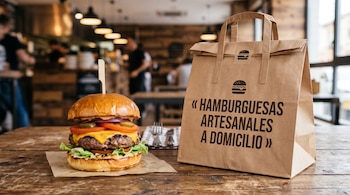 Una hamburguesa con queso, lechuga y tomate junto a una bolsa de papel marrón sobre una mesa de madera con un salón desenfocado al fondo.