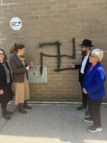 Cuatro personas observan una esvástica negra pintada con aerosol en la pared de ladrillo de una sinagoga; se ve una placa y un cartel de vigilancia