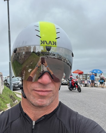 Un hombre con casco blanco y amarillo, gafas reflectantes y camiseta deportiva oscura toma una selfie. Sus gafas reflejan una calle con vehículos y la costa