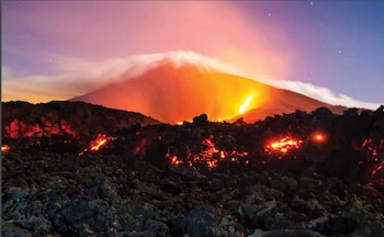 Vista nocturna del Volcán de Fuego en erupción con lava incandescente fluyendo por sus laderas y un penacho de ceniza en la cima, bajo un cielo estrellado