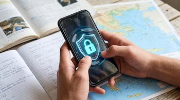 Dos manos sostienen un teléfono móvil con un icono de candado iluminado sobre un cuaderno escolar y un mapa de Grecia.