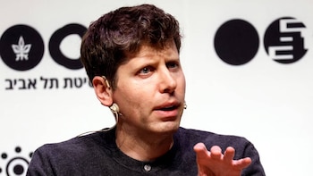 Primer plano de Sam Altman con cabello castaño oscuro, ojos azules y piel clara, usando un auricular, hablando frente a un fondo blanco con texto en hebreo