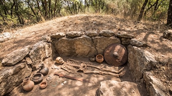 Tumba abierta en tierra con paredes de piedra, un esqueleto humano, un escudo, una espada, varias vasijas y otros objetos metálicos.