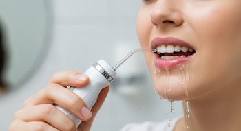 Primer plano de una mujer utilizando un irrigador dental blanco, con la boquilla insertada en su boca y agua goteando por la barbilla.