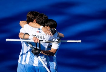 Tres jugadores vestidos con la camiseta argentina se abrazan sobre un campo azul sujetando palos de hockey