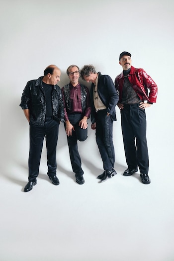 Cuatro hombres, los integrantes de Cuarteto de Nos, posan de pie frente a una pared blanca. Visten chaquetas y pantalones oscuros