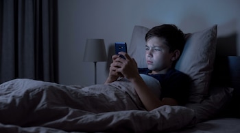Niño acostado en una cama a oscuras, usa un celular que ilumina su rostro. Se observan almohadas, una manta, una mesita de noche y parte de una lámpara.