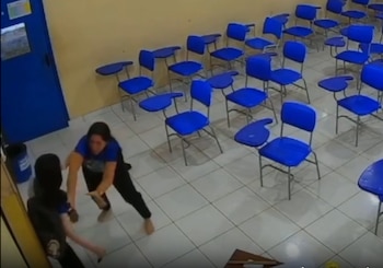 Dos personas de pie junto a una puerta azul dentro de un aula con pupitres de plástico azul