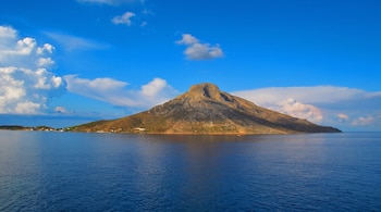 Una gran isla montañosa y rocosa se eleva del mar bajo un cielo azul con nubes blancas. Se observa una franja costera con casas a la izquierda.