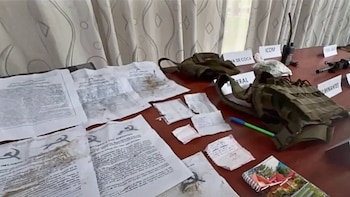 Varios documentos con un martillo y una hoz, papeles pequeños, un chaleco táctico, una radio, partes de un fusil, bolsas con contenido, un cuaderno y un bolígrafo sobre una mesa