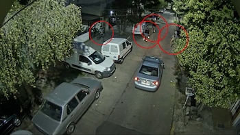 Vista aérea nocturna de una calle con varios autos y furgonetas estacionados, y personas circulando en bicicletas. Árboles con follaje verde a los lados