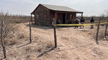 Cabaña de madera con cinta amarilla de la policía y varios agentes uniformados en un terreno árido.