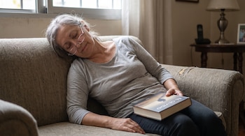 Una mujer de cabello gris con gafas duerme en un sofá marrón; tiene un libro sobre su regazo, y su cabeza está inclinada hacia un lado.