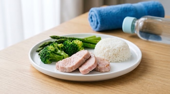 Plato blanco con filetes de cerdo, brócoli, espárragos y arroz blanco en una mesa de madera. Al fondo, una toalla azul enrollada y una botella de agua.