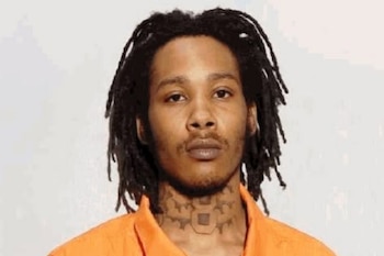 Fotografía policial de Eljay Crisp-Carr, un hombre con rastas y barba incipiente, usando una camisa naranja, mirando directamente a la cámara con un fondo liso