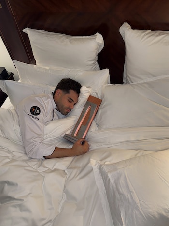 Un hombre con chaqueta blanca de cocinero descansa en una cama con sábanas blancas mientras sujeta un trofeo con forma de cuchillo