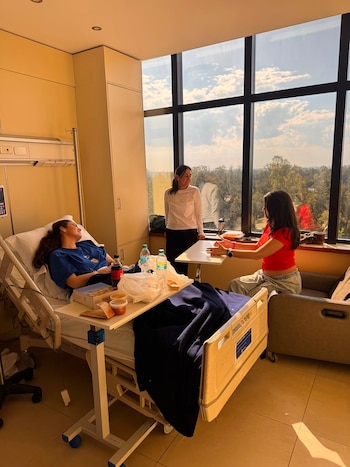 Mujer recostada en cama de hospital con bata de paciente azul; dos mujeres de pie y sentada a su alrededor; mesa con comida; ventana grande con vista a árboles y cielo