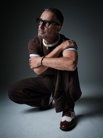 Jorge Drexler, hombre canoso con barba y gafas, agachado y mirando a la izquierda. Viste camiseta marrón, pantalones a rayas y zapatos bicolor