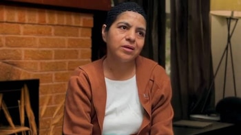 Una mujer con gorro negro y chaqueta marrón permanece sentada ante una chimenea de ladrillo