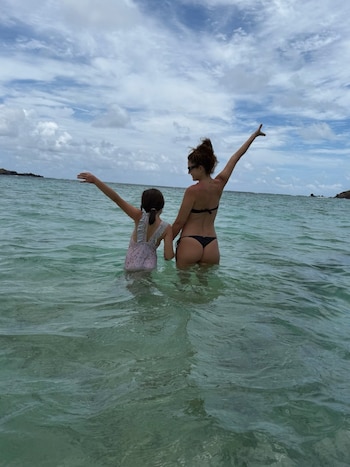 Mujer con bikini negro y niña con traje de baño rosa de espaldas en el mar con los brazos en alto. Cielo nublado y rocas en la distancia