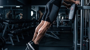 Persona con ropa deportiva entrena las pantorrillas en una máquina de gimnasio, con pesas y otras máquinas desenfocadas al fondo.