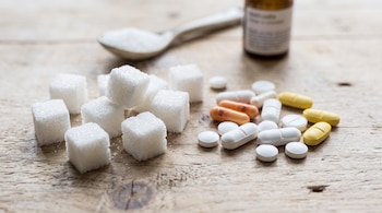 Terrones de azúcar blanco, pastillas de diversos colores, una cuchara llena de azúcar y un frasco sobre una mesa de madera.