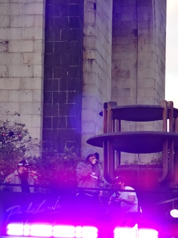 Un cantante con sudadera de rayas sostiene un micrófono en una plataforma iluminada de color violeta junto a una pared de piedra