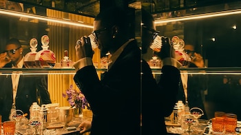 Hombre de perfil bebiendo de un vaso de whisky, reflejado en un espejo, junto a una barra con botellas decoradas, cocteleras, copas y flores