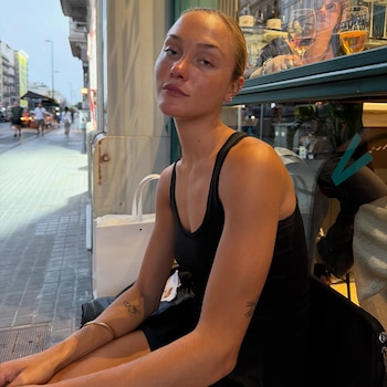 Mujer joven con top negro de tirantes sentada en una silla. Presenta tatuajes en los brazos. Al fondo hay una ventana, una calle con edificios, coches y personas