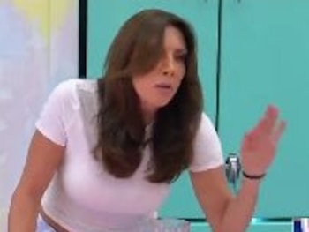 Mujer con camiseta blanca y pantalón corto a cuadros detrás de una mesa con un vaso de líquido oscuro, fondo de estudio azul y amarillo, texto en pantalla