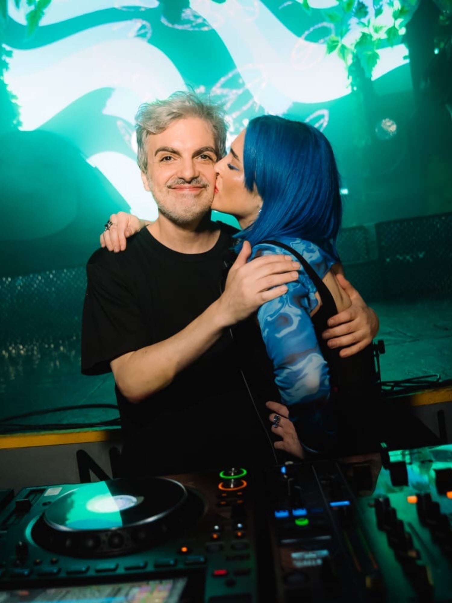 Cintia besa en la mejilla al músico Ale Sergi mientras posan abrazados detrás de una consola de DJ en un evento con iluminación verde.