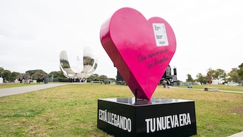 Escultura de corazón rosa y base negra con texto, situada en un parque de césped. Al fondo se observa la Floralis Genérica y arboledas