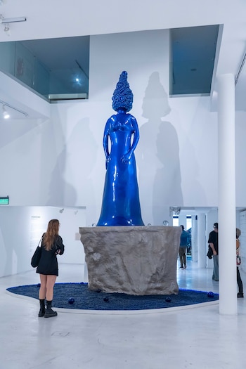Escultura azul brillante de figura humana con cabeza texturizada sobre pedestal gris en una sala de museo blanca, con una persona de espaldas observándola