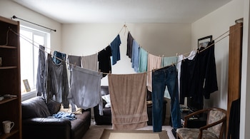 Cuerda tensada con ropa tendida incluye camisas, pantalones y toallas sujetas con pinzas; se aprecian dos sofás y una estantería de madera.