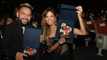 Anna Carina Copello y Jesús 'El Viejo' Rodríguez sonríen mientras sostienen premios dorados con el mapa de Perú, sentados en un auditorio