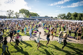 Multitud de personas haciendo ejercicio o bailando al aire libre en un día soleado, sobre césped verde, con carpas blancas y árboles en el fondo