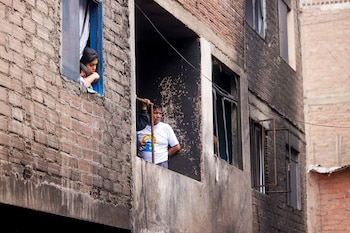 Fachada de edificio de ladrillo con paredes oscuras y ventanas rotas. Una mujer y un niño miran por una ventana, y un hombre por otra, sosteniendo un palo