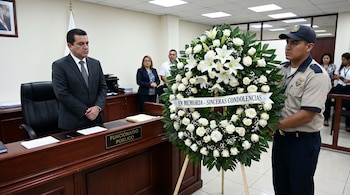 Un hombre en traje mira una corona fúnebre grande de rosas y lirios blancos, sostenida por un hombre uniformado en una oficina. Otros observan al fondo.
