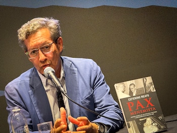 Hombre de mediana edad con gafas y chaqueta azul habla en un micrófono, gesticulando con las manos. Un libro "PAX MENEMISTA" de Ceferino Reato y una botella de agua están a su lado